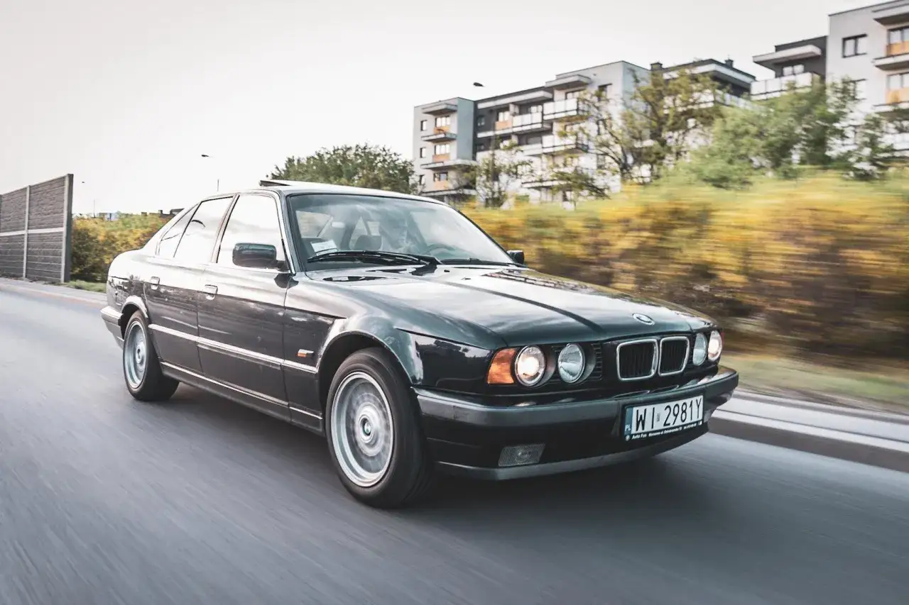 Ciemne BMW E34 w ruchu, z rozmytym tłem budynków i drzew. Klasyk na drodze.