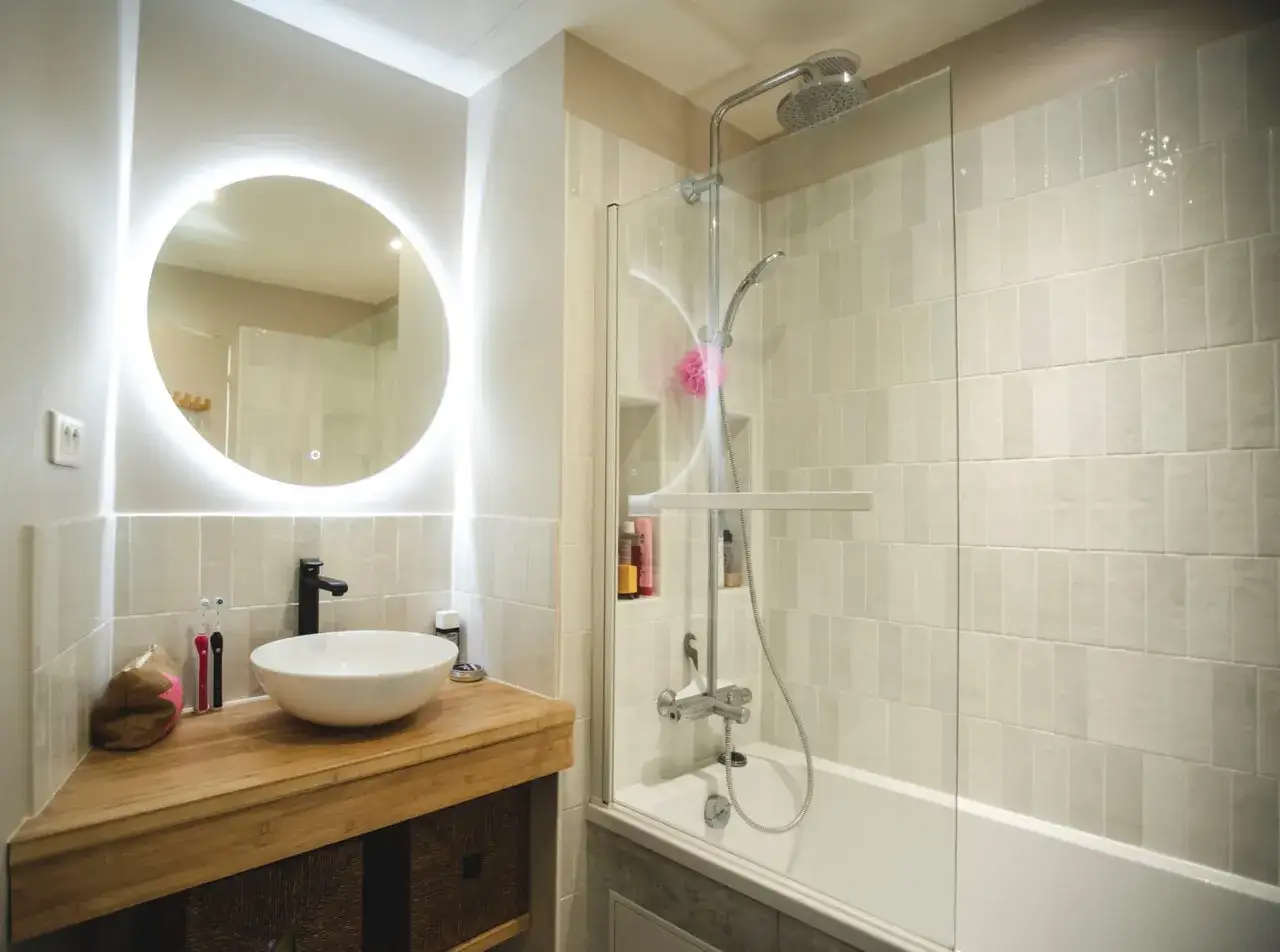 Salle de bain sans fen&ecirc;tre, avec un grand miroir rond lumineux, une vasque moderne et une douche/baignoire.