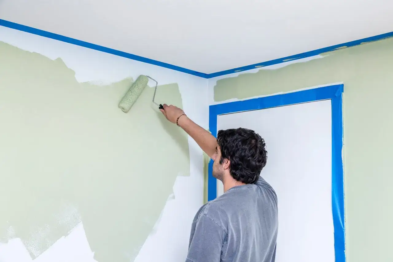 Mann streicht mit einer Farbrolle eine Wand hellgrün. Blaue Malerkrepp schützt die Decke und den Türrahmen.