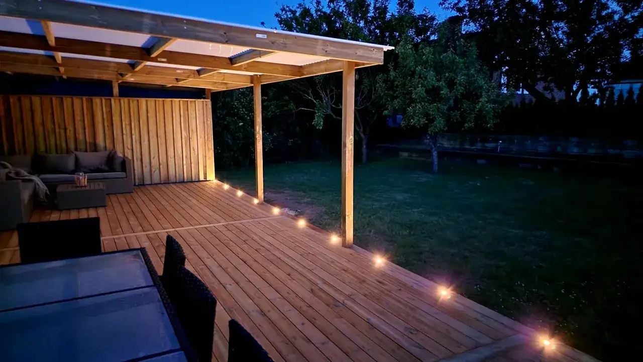 En mysig altan med en pergola som har plasttak. Trädäck med belysning längs kanten.
