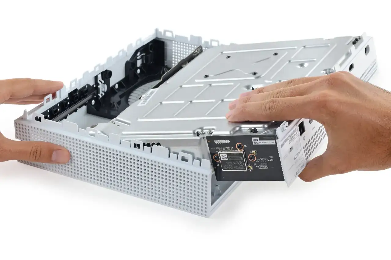 Wymiana dysku Xbox One S. Dłonie wyjmują białą płytkę z wnętrza konsoli, odsłaniając jej mechanizm.