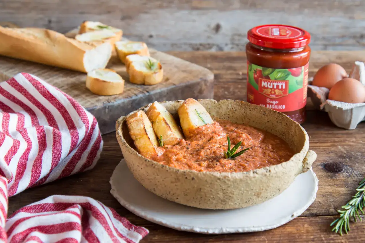 Un piatto rustico di uova al pomodoro, guarnito con rosmarino e accompagnato da crostini di pane. Sullo sfondo, un barattolo di sugo Mutti e uova fresche.