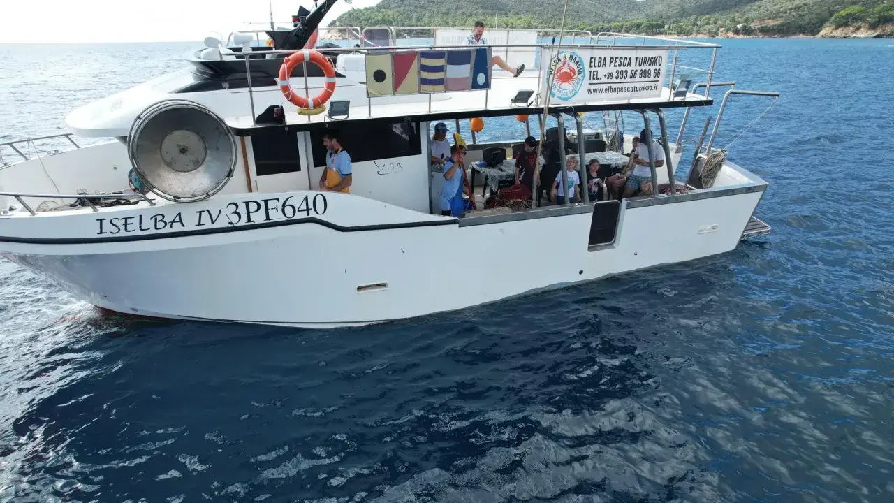 Barca bianca in navigazione, con persone a bordo per un'esperienza di pescaturismo. Sul fianco la scritta 