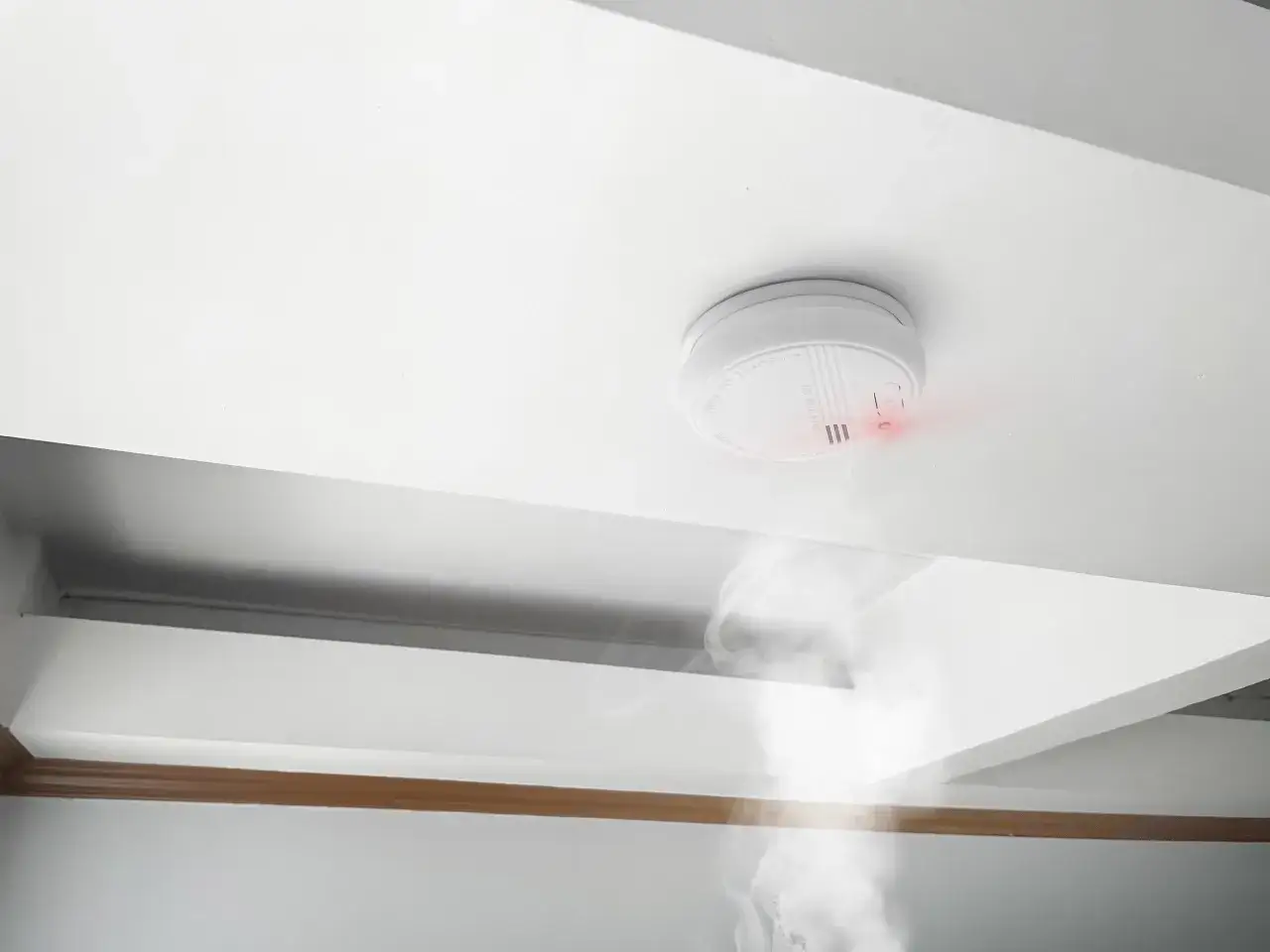 Detector de humo blanco en techo, emitiendo luz roja y humo. Existen varios tipos de detectores de humo para tu seguridad.