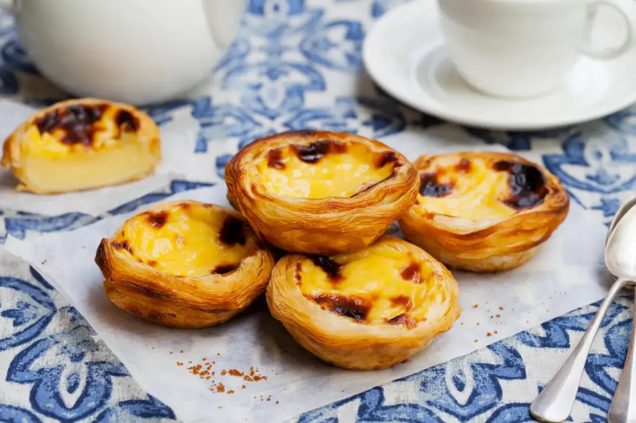 Cztery pyszne, lekko przypieczone pastel de nata na papierze, obok filiżanki kawy.