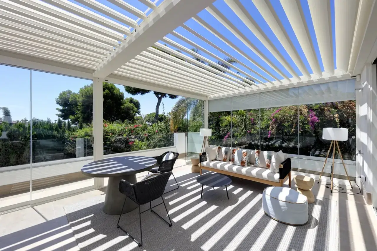 Une veranda bioclimatique, c'est quoi ? Un espace de vie lumineux avec salon et table, entouré de verdure, sous une pergola ajourée.