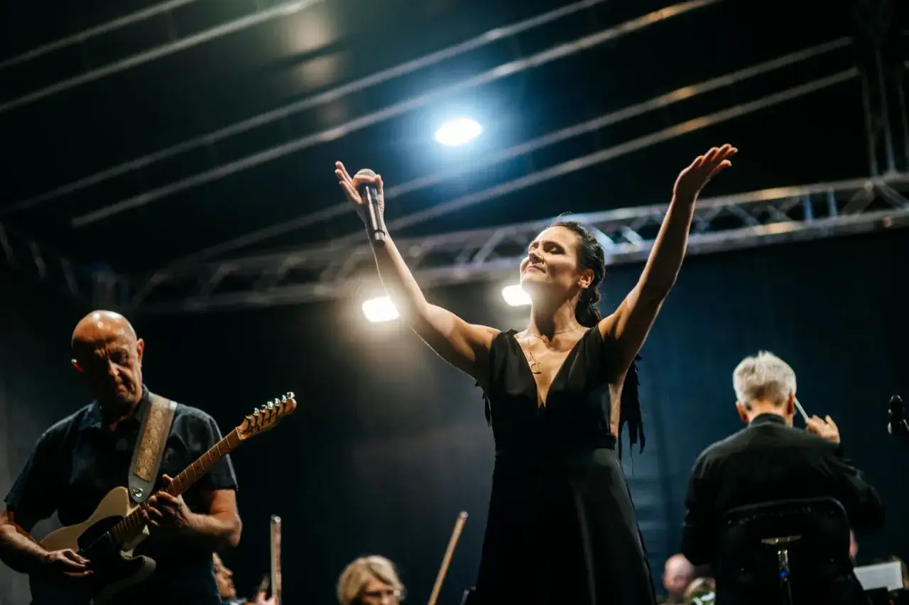 Wokalistka z podniesionymi rękami śpiewa, obok gitarzysta i dyrygent. Cały zespół z korą tworzy magiczną atmosferę.