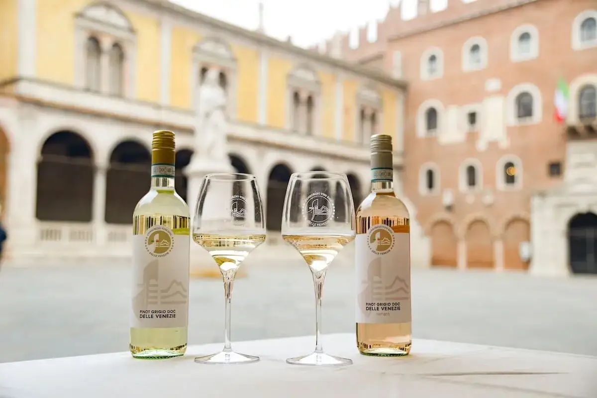Due bottiglie e due calici di Pinot Grigio Delle Venezie, uno bianco e uno ramato, su un tavolo con un edificio storico sullo sfondo. Perfetto sia fermo che frizzante.