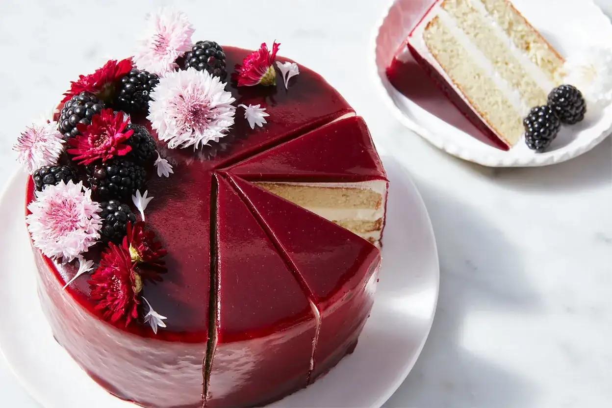 Ein Kuchen mit gl&auml;nzender, roter mirror glaze, dekoriert mit Brombeeren und zarten Bl&uuml;ten. Ein St&uuml;ck liegt auf einem Teller.