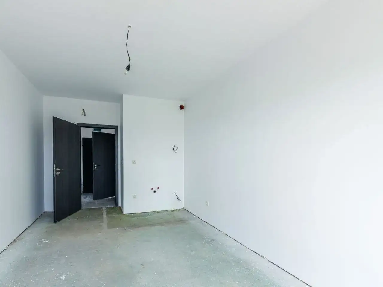 Puste pomieszczenie w stanie deweloperskim. Widać betonową podłogę, białe ściany, otwarte drzwi i luźny kabel z żarówką.