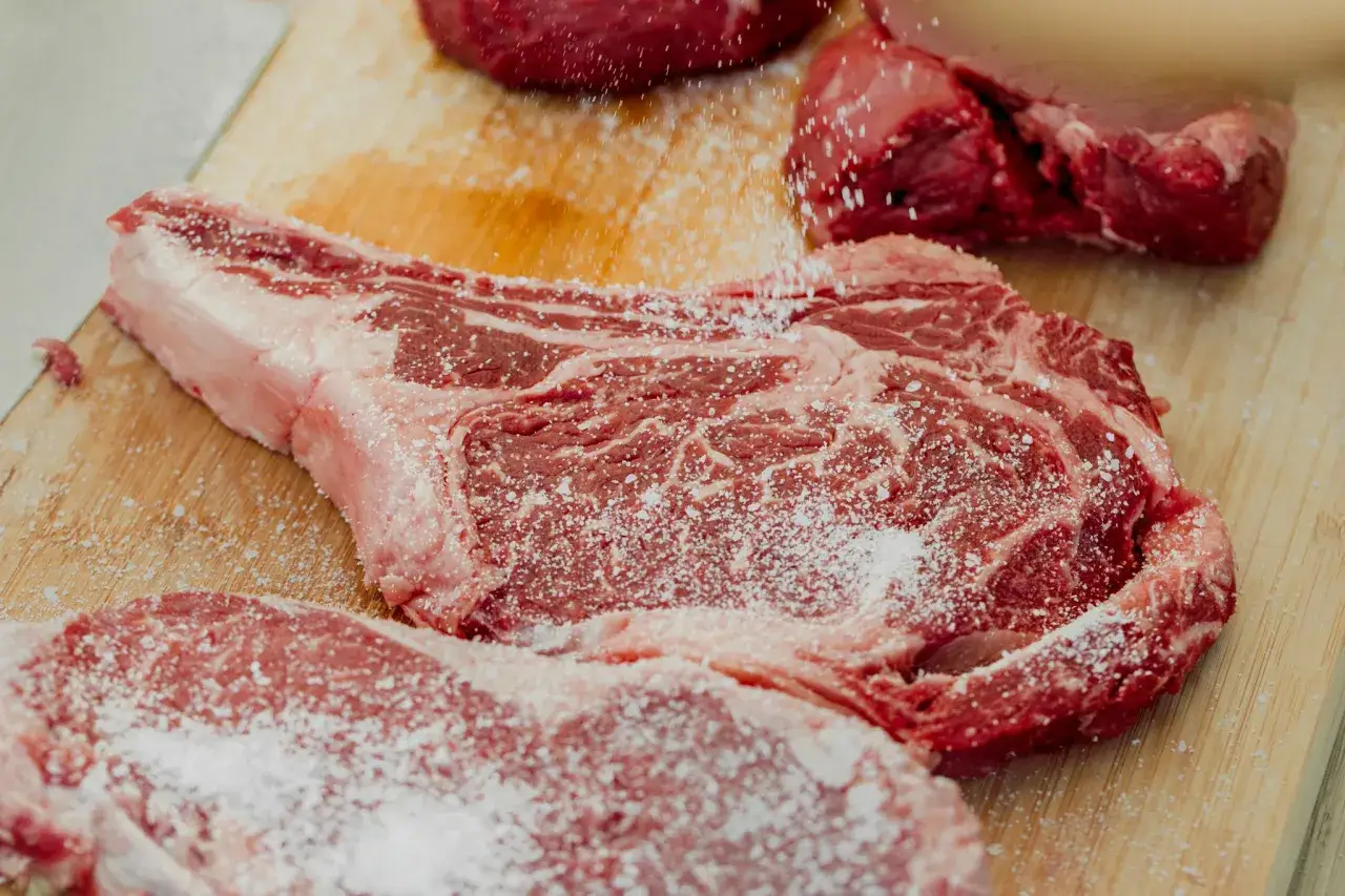 Gros morceaux de viande rouge, dont une c&ocirc;te de b&oelig;uf, sont assaisonn&eacute;s de gros sel. Le persillage de la viande est bien visible, promettant une cuisson savoureuse.