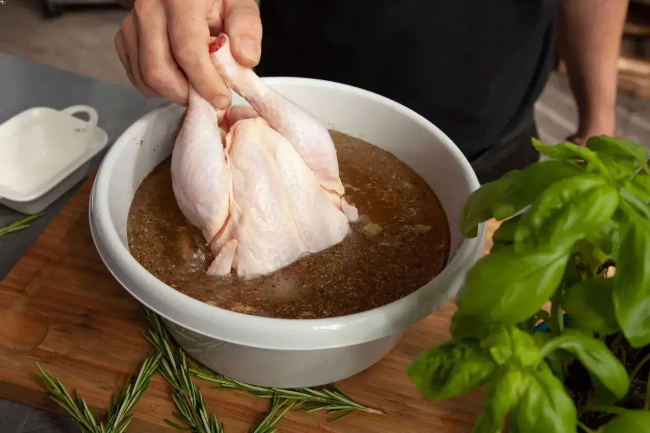 Ein rohes H&auml;hnchen wird in eine Sch&uuml;ssel mit einer w&uuml;rzigen Fl&uuml;ssigkeit getaucht, um Fleisch in Salzlake einzulegen. Frischer Basilikum und Rosmarin liegen daneben.