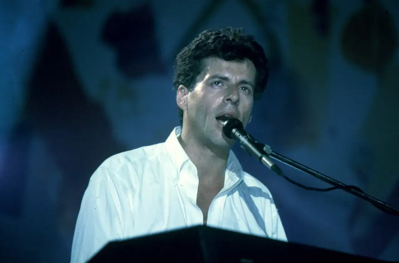 Claudio Baglioni, icona della musica italiana, canta al microfono durante un concerto. La sua biografia &egrave; un viaggio nella melodia.