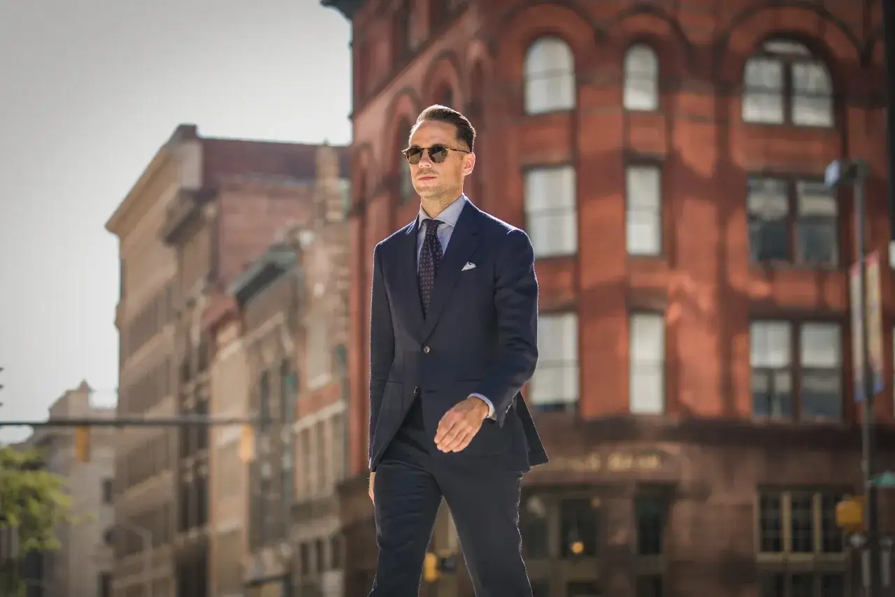 A stylish man demonstrates how to wear a blue suit with a patterned tie and pocket square, walking confidently past brick buildings.