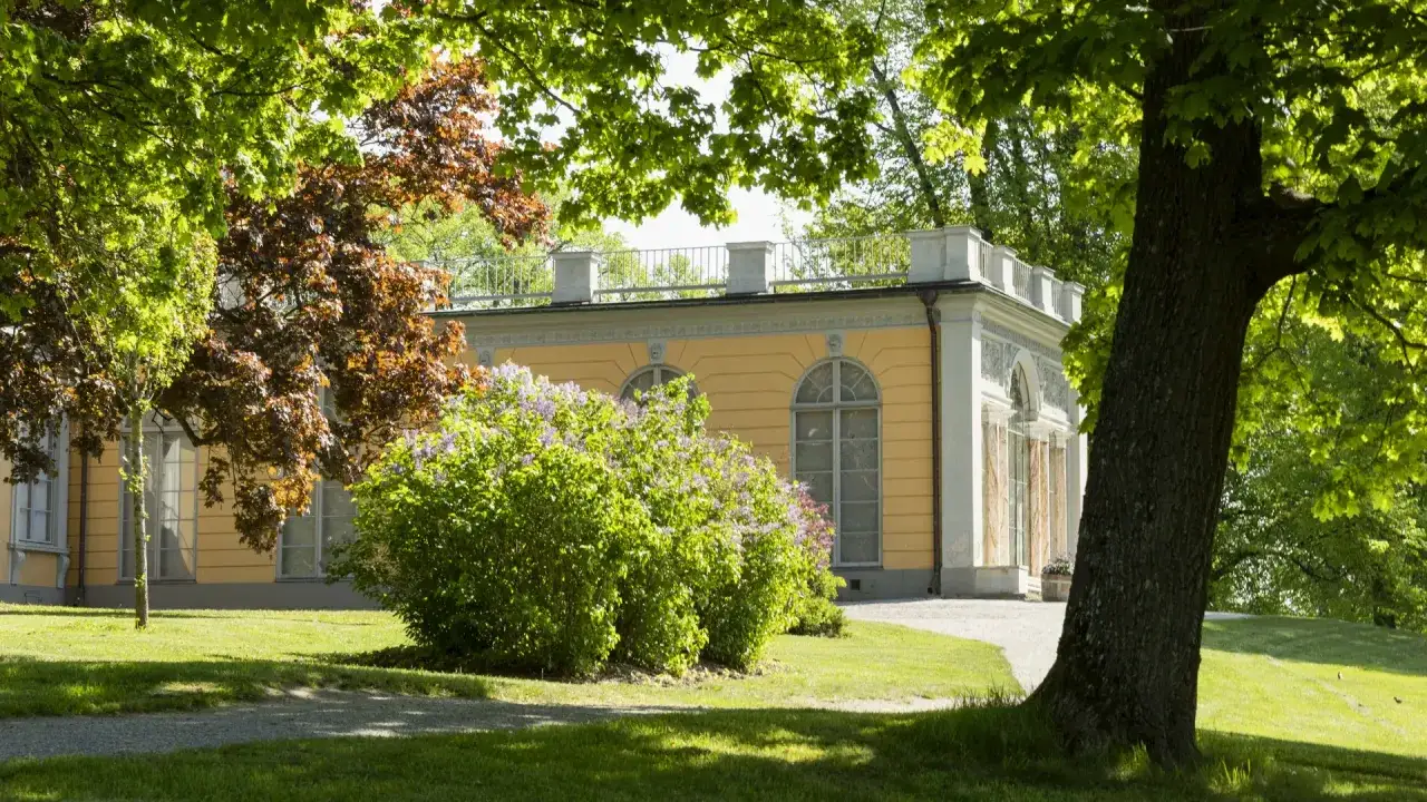 En gul byggnad med vita detaljer i Haga parken, omgiven av grönskande träd och buskar.
