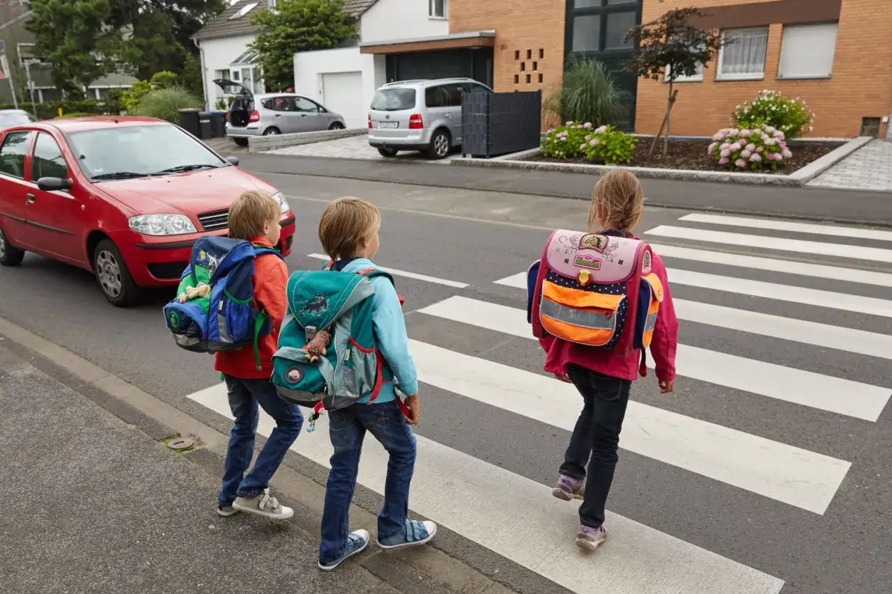 Drei Schulkinder mit Rucks&auml;cken &uuml;berqueren vorsichtig die Stra&szlig;e. Ein rotes Auto wartet. Ein sicherer Schulweg f&uuml;r die Grundschule.