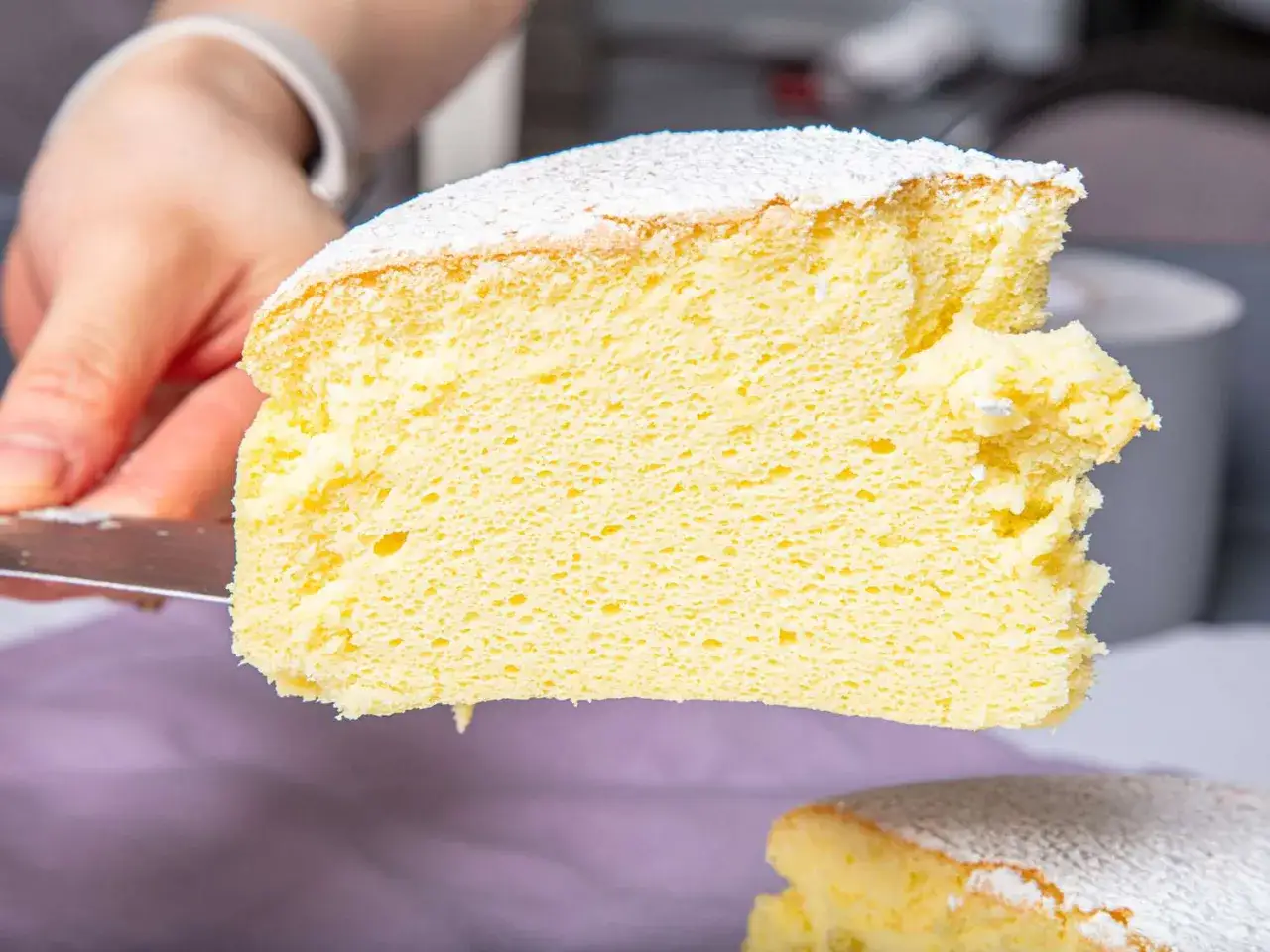 Une main coupe une tranche a&eacute;rienne de cheesecake japonais saupoudr&eacute; de sucre glace. Sa texture l&eacute;g&egrave;re et moelleuse invite &agrave; la d&eacute;gustation.