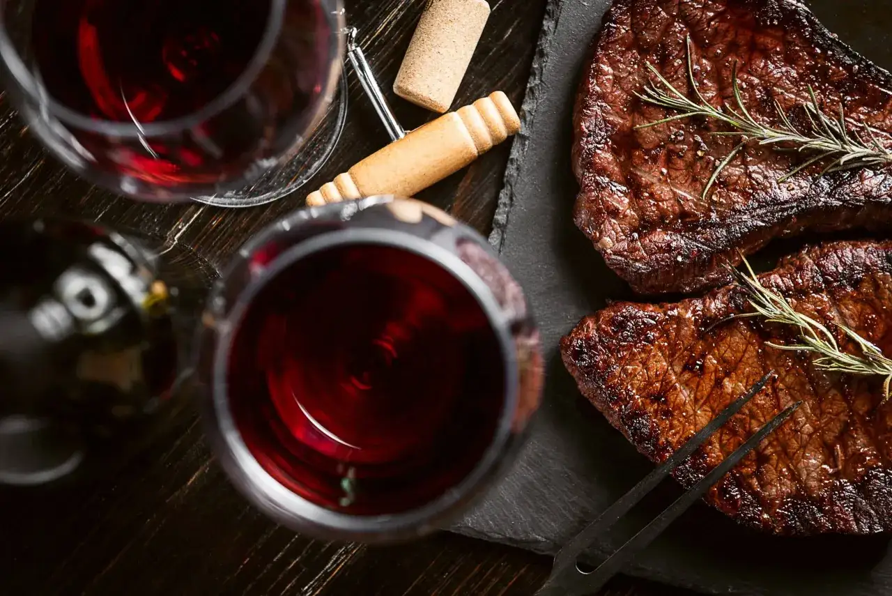 Deux verres de vin rouge, une bouteille, un tire-bouchon et deux savoureuses viandes rouges grill&eacute;es garnies de romarin.