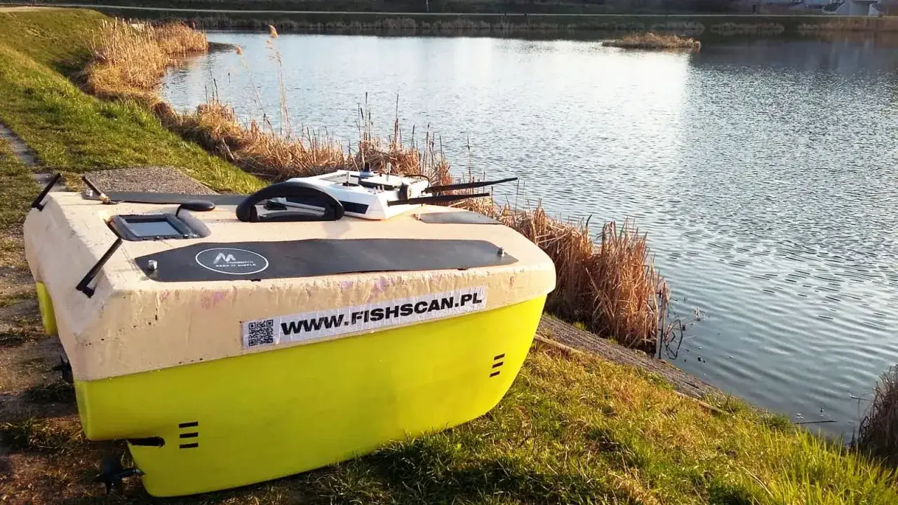 Żółto-beżowy pojazd wodny, prawdopodobnie do badań zbiornika, stoi na brzegu. Na burcie widnieje napis WWW.FISHSCAN.PL.