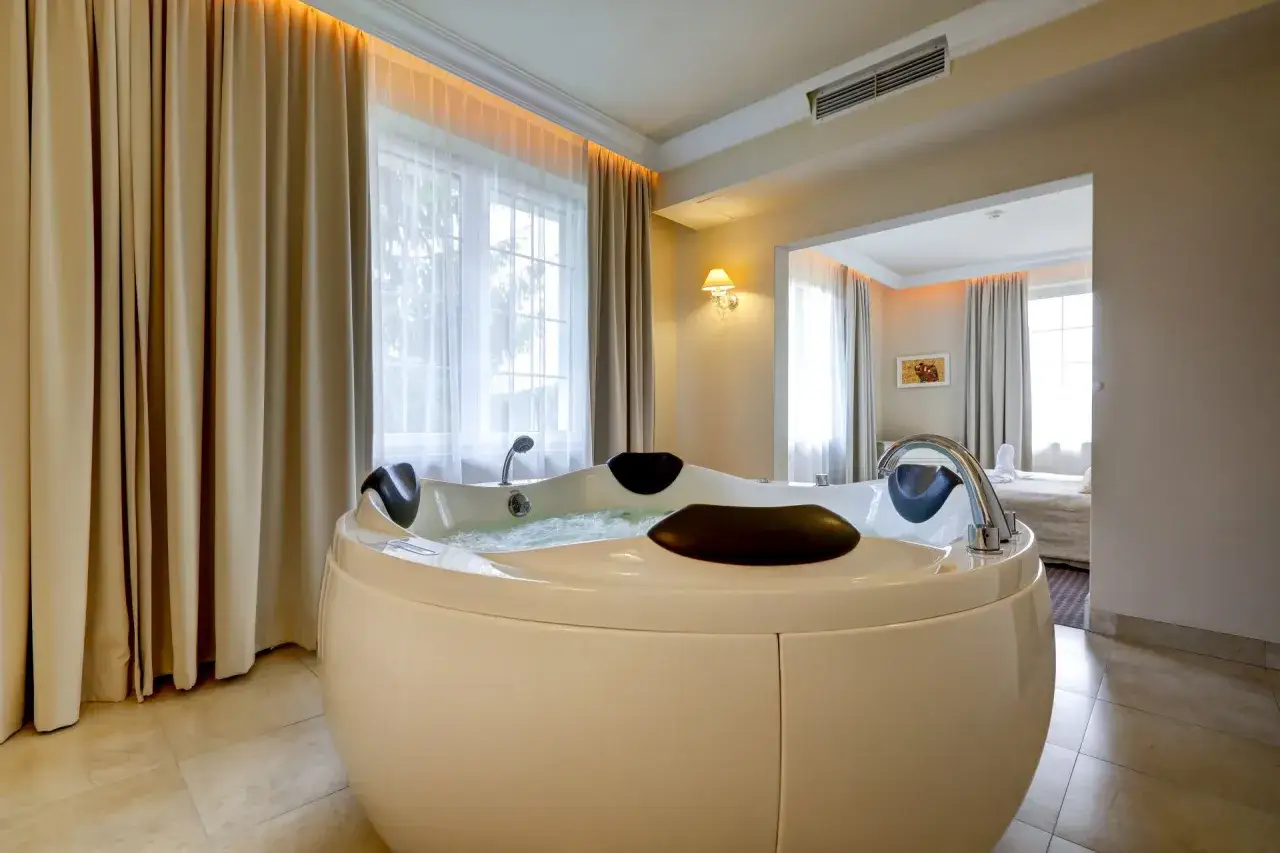 Nowoczesny projekt pokoju hotelowego z okrągłą wanną z hydromasażem. Eleganckie zasłony i subtelne oświetlenie tworzą przytulną atmosferę.