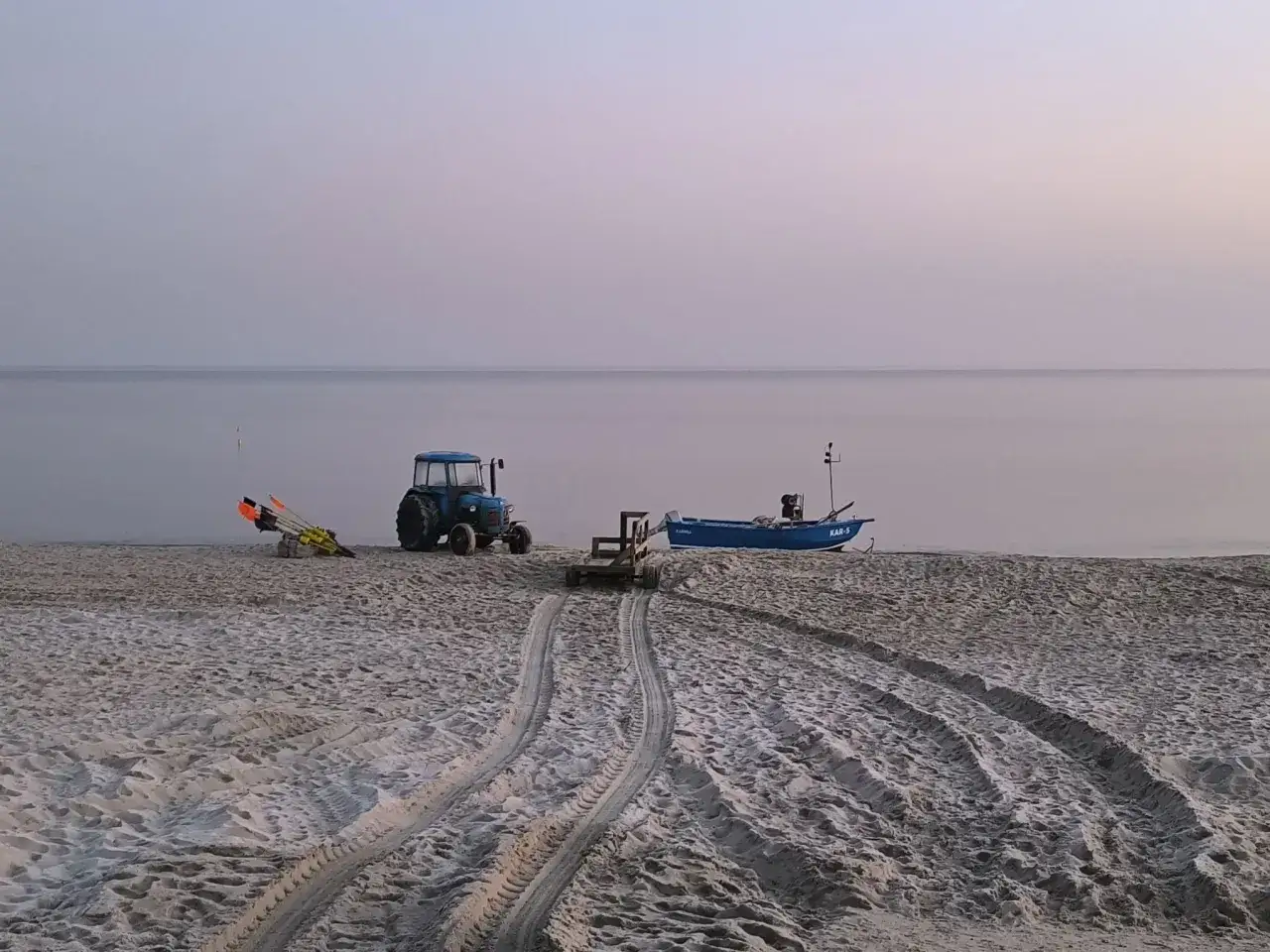 Niebieski traktor i łódź na plaży w Karwi. Ślady opon na piasku prowadzą do morza.