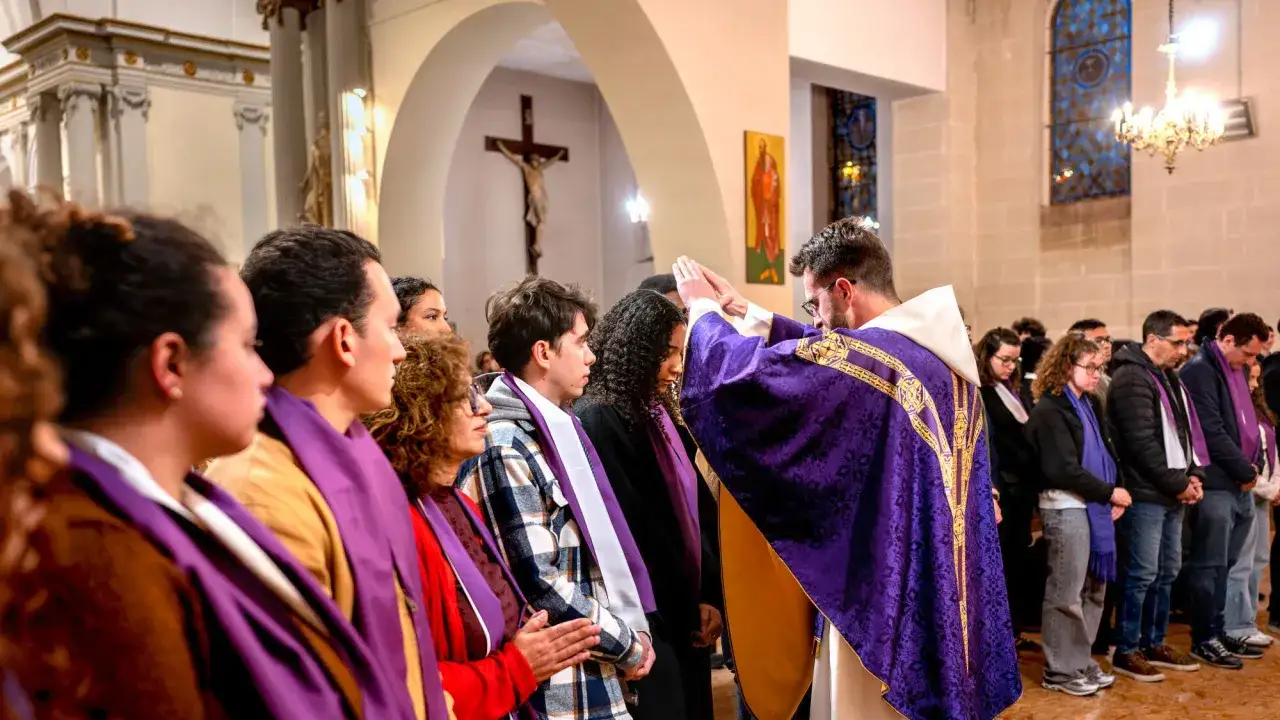 Un pr&ecirc;tre b&eacute;nit des fid&egrave;les en pr&eacute;paration au bapt&ecirc;me. Les paroissiens, v&ecirc;tus de ch&acirc;les violets, &eacute;coutent attentivement dans une &eacute;glise.