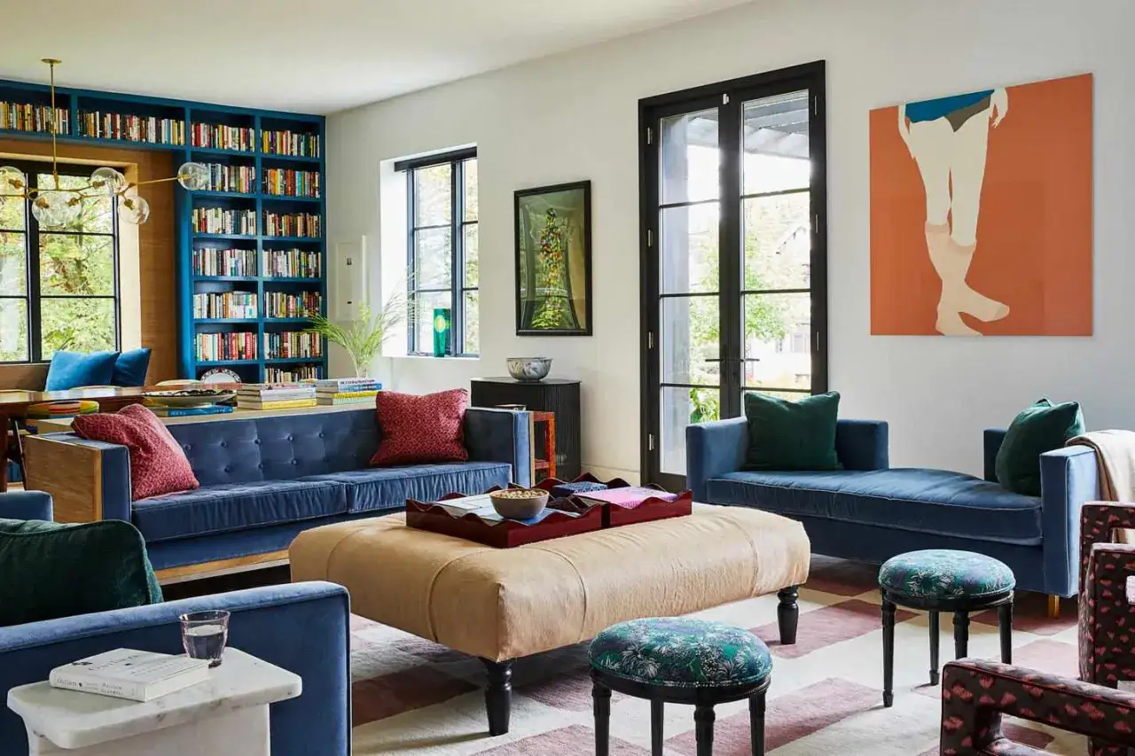 Gemütliches Wohnzimmer mit **moderner Einrichtung**: blaue Sofas, ein großer beiger Hocker, Bücherregale und Kunst an der Wand.