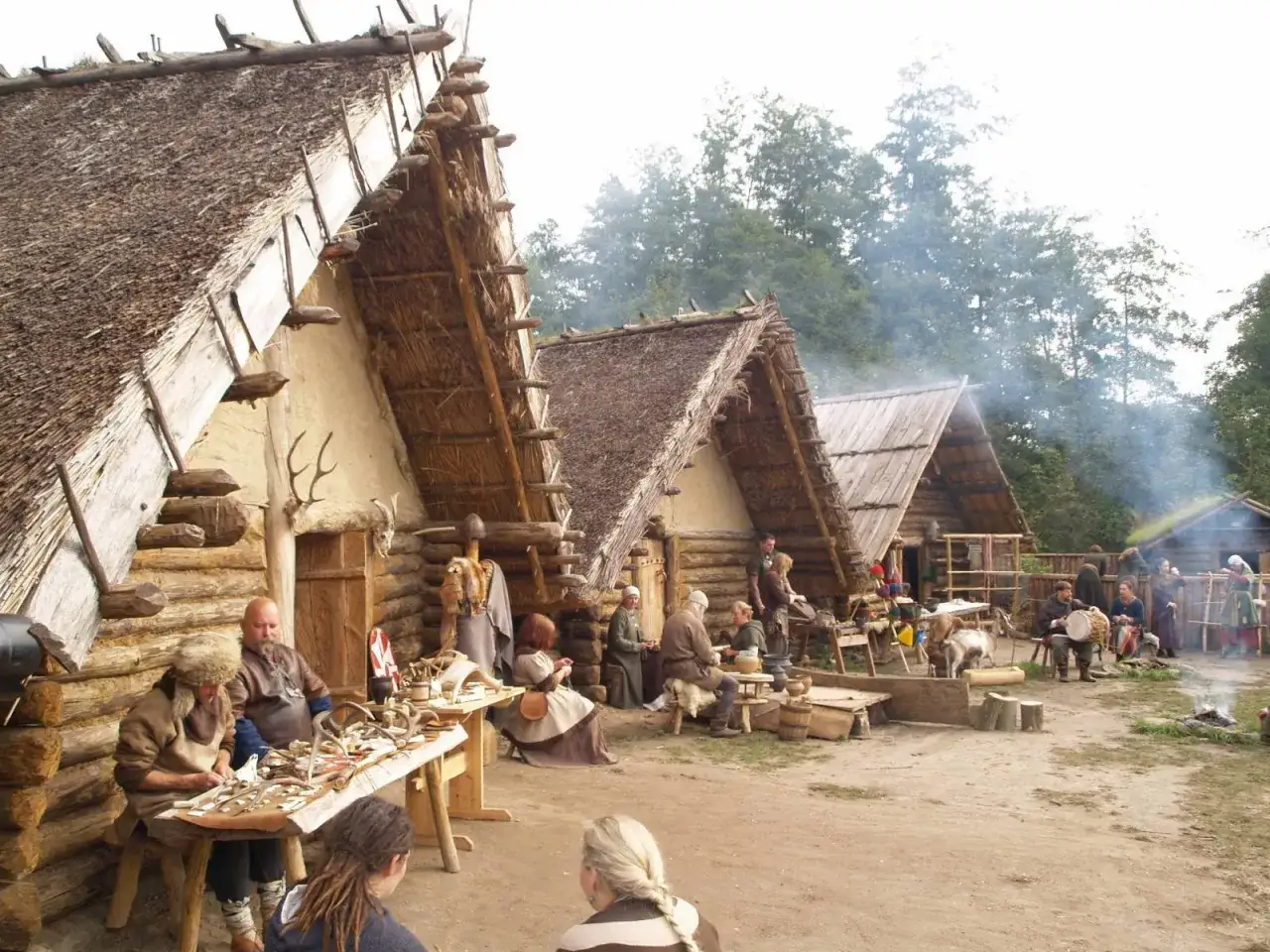 Odwzorowanie osady Biskupin z ludźmi w strojach historycznych, pracującymi i odpoczywającymi przy drewnianych domach krytych strzechą.