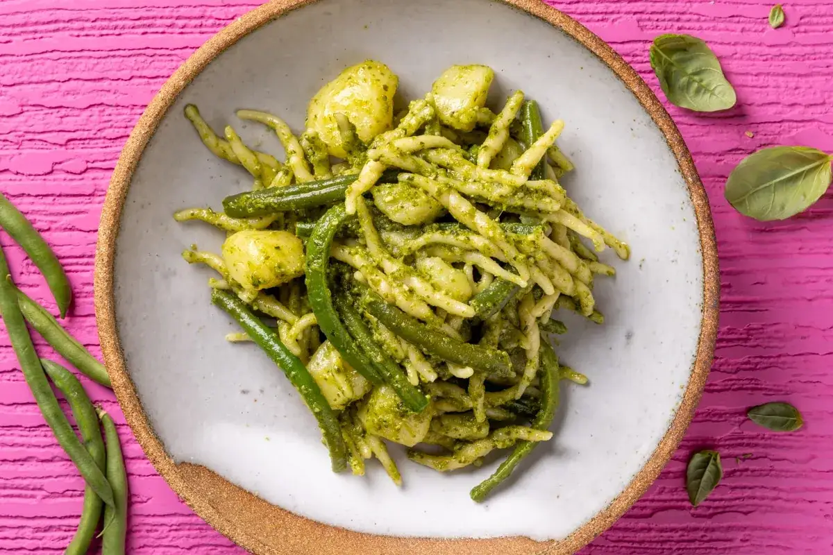 Un piatto di pasta al pesto con fagiolini e patate, su uno sfondo rosa vibrante. Fresco e invitante.