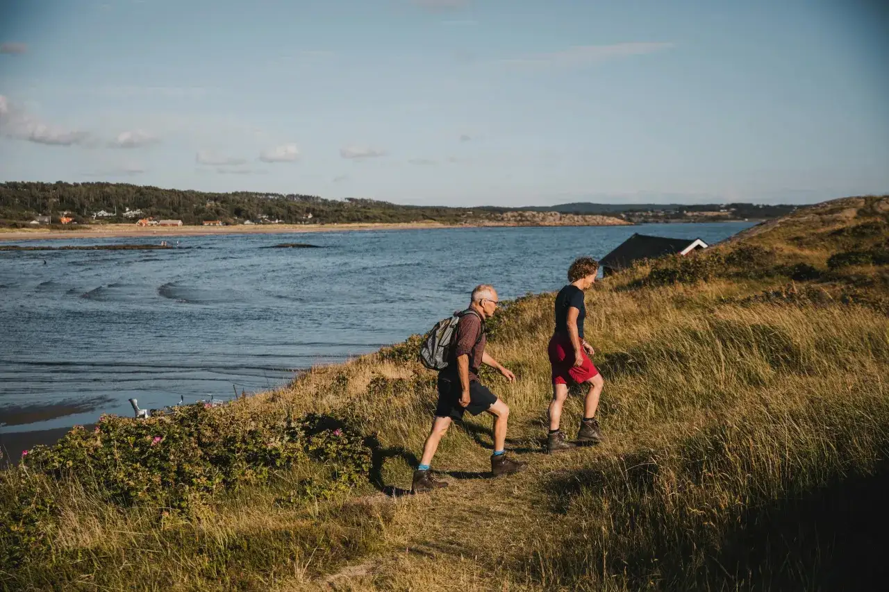 Två personer vandrar längs en gräsbevuxen kuststig nära Rosendals camping. Havet glittrar i bakgrunden.