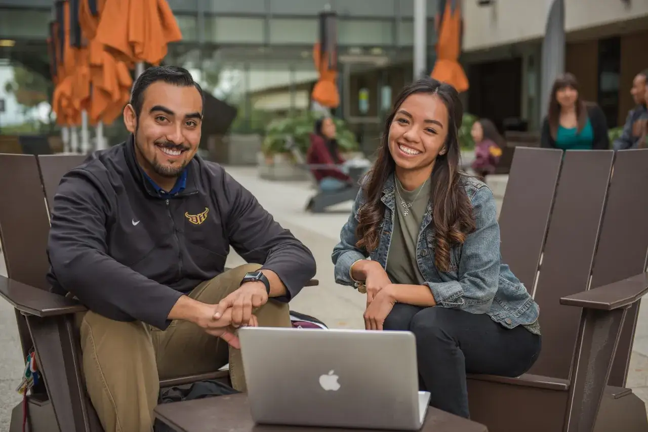 Two smiling students, likely pursuing an MPA degree in California, share a laptop outdoors.