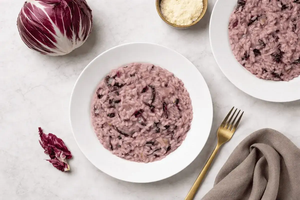Un cremoso risotto al radicchio rosso, con un cespo intero e una foglia decorativa. Servito in piatti bianchi con forchetta dorata.