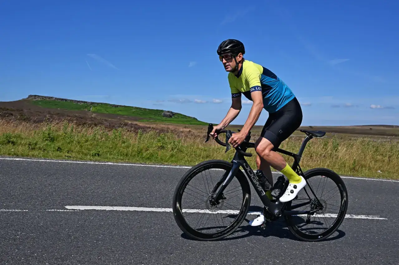 A cyclist in a yellow and blue jersey and black shorts is biking 50 miles a week on a paved road through a grassy, hilly landscape under a clear blue sky.