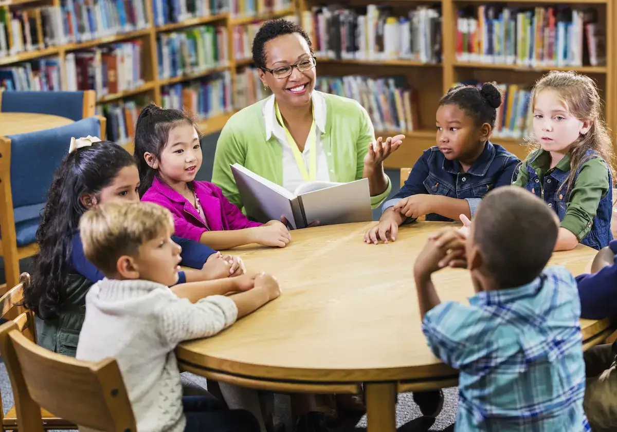 Lehrerin liest Kindern im B&uuml;cherregal vor. Die Kinder lauschen gespannt, was das Lesen f&uuml;r sie bedeutet.