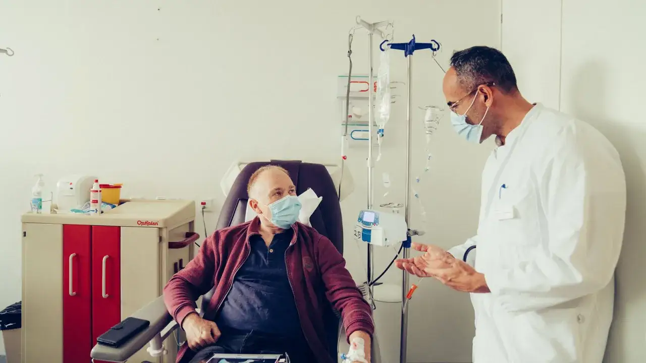Arzt spricht mit Patient während adjuvanter Chemotherapie. Der Patient sitzt in einem Sessel, Infusionsständer und medizinische Geräte sind sichtbar.