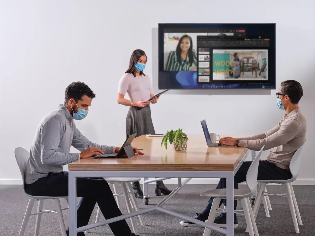 Zwei M&auml;nner arbeiten am Laptop, eine Frau steht daneben. Auf dem Bildschirm ist eine Videokonferenz zu sehen &ndash; ein Beispiel f&uuml;r hybrides Arbeiten.