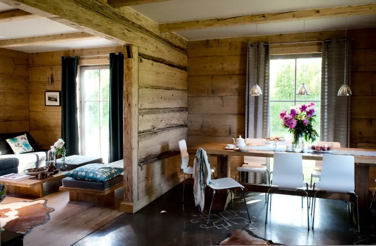 Drewniane ściany i belki tworzą przytulną atmosferę w stylu modern farmhouse. W salonie sofa i ława, w jadalni stół z białymi krzesłami.