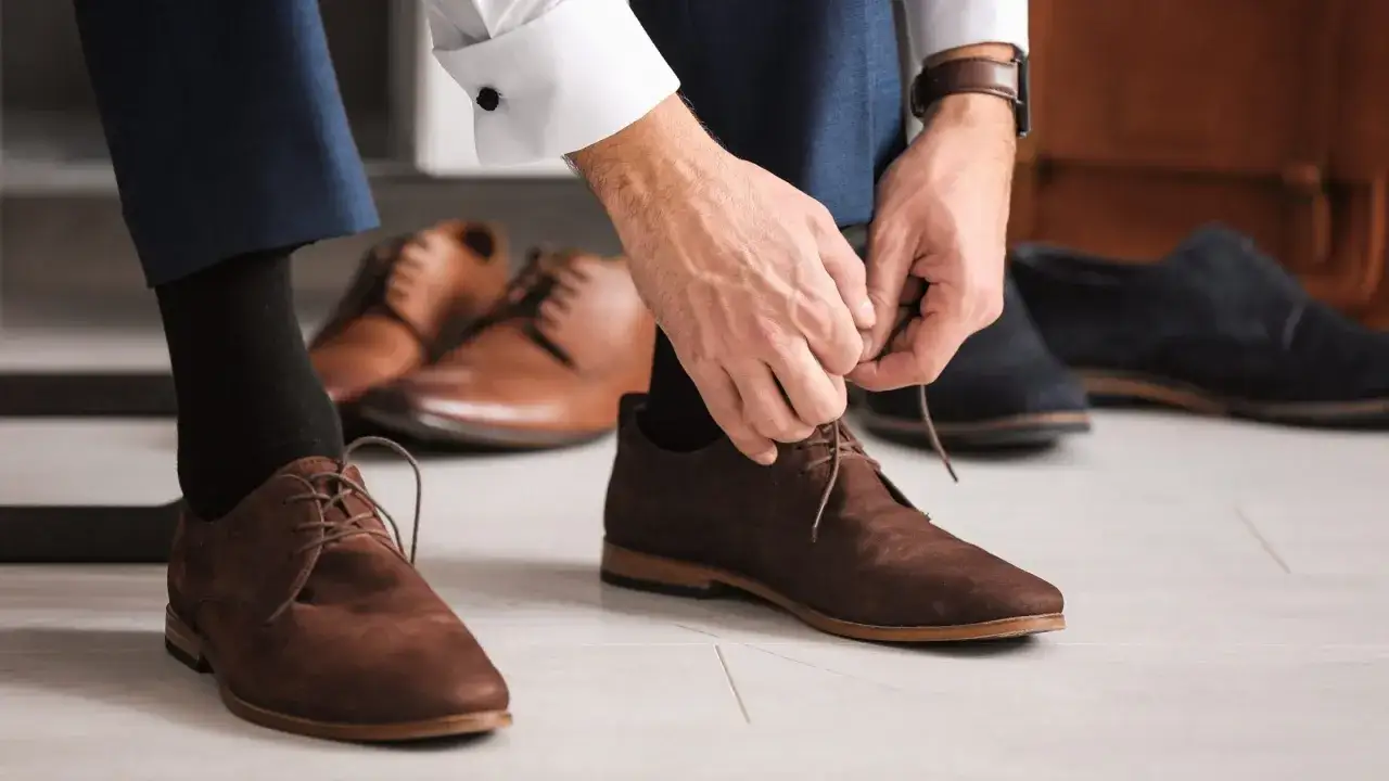 Mann schnürt braune Wildlederschuhe, die perfekt zu seinem Anzug passen. Mehrere andere Anzugschuhe liegen bereit.
