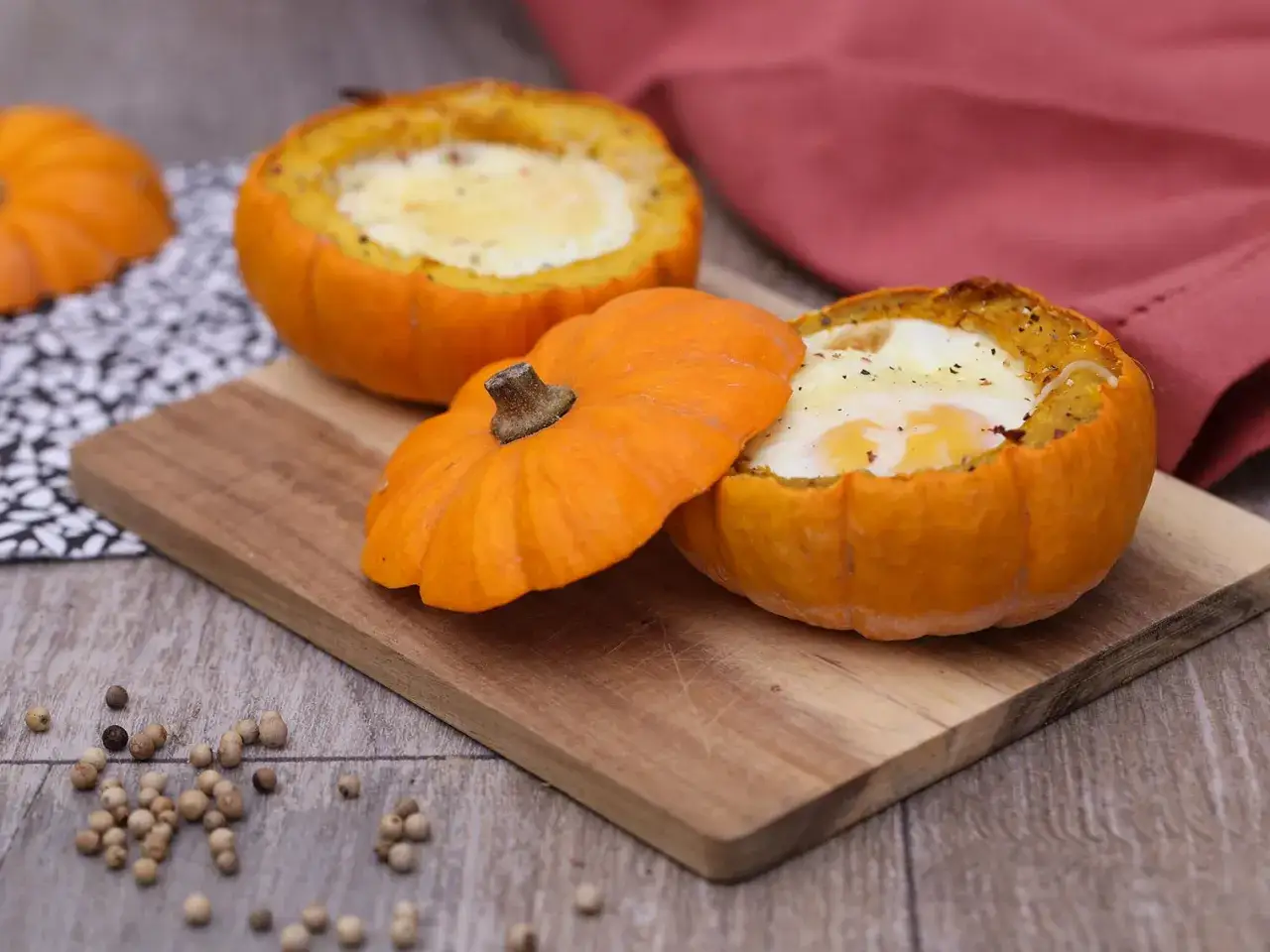Œufs cuits dans des little jack courges, servis sur une planche en bois avec des grains de poivre.