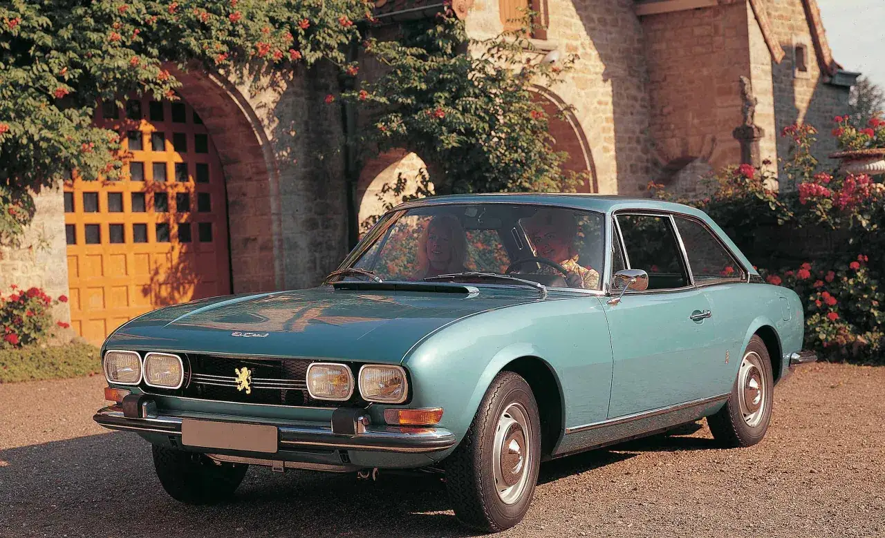 Elegancki, błękitny Peugeot 504 Coupe z parą pasażerów przed zabytkowym budynkiem z kamienia i kwitnącymi krzewami.
