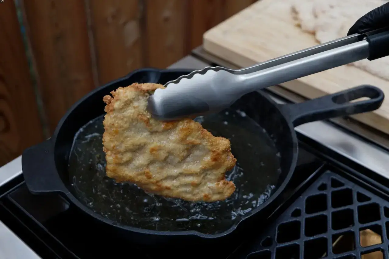 Ein paniertes Schnitzel wird mit einer Zange in heißem Öl in einer Gusseisenpfanne gebraten. Perfekt für ein schnelles schnitzel rezept.
