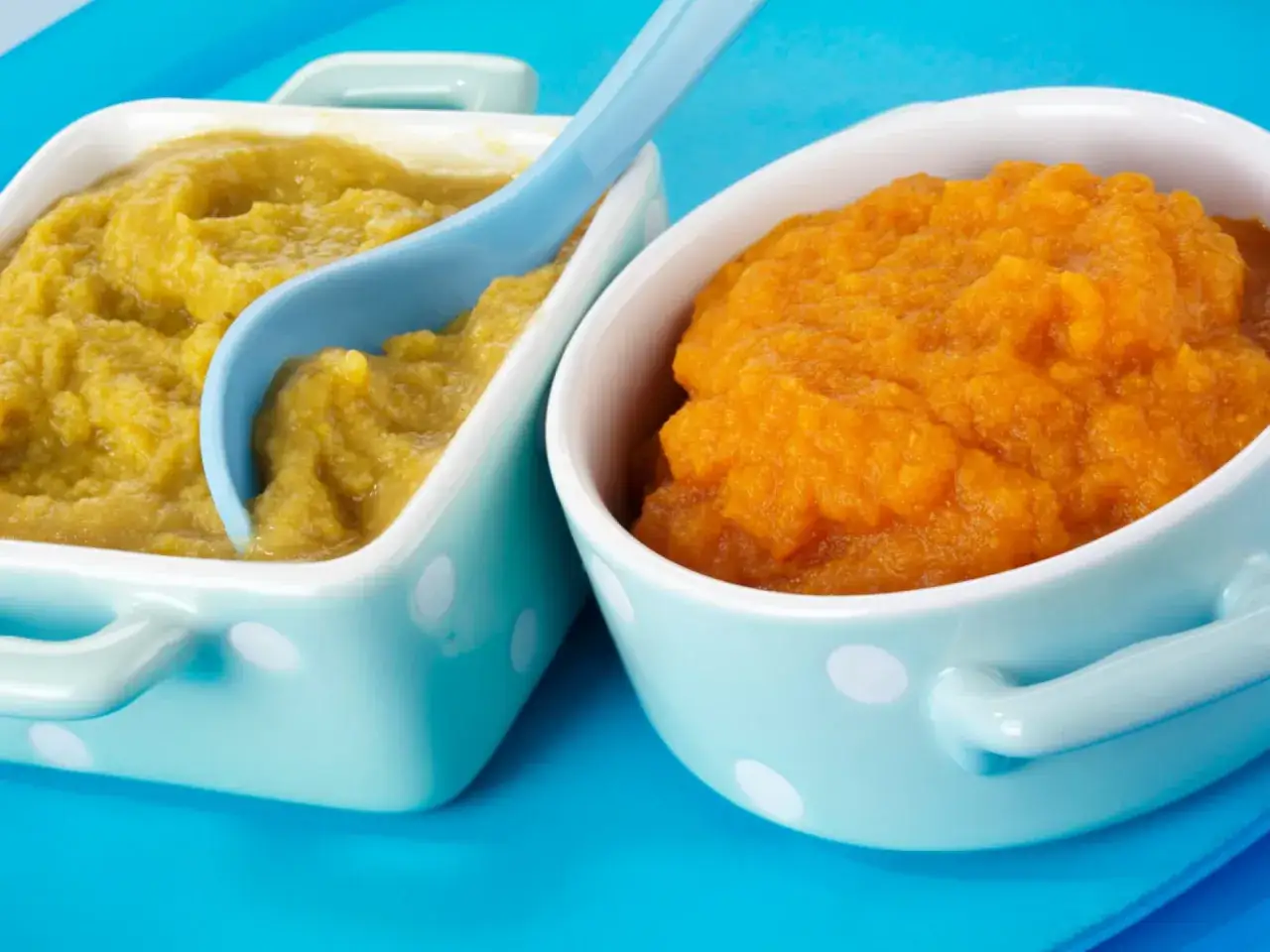 Zwei Schalen mit Babybrei: eine gelbgr&uuml;ne, eine orangefarbene. Ein blauer L&ouml;ffel liegt in der gelbgr&uuml;nen Portion. Perfekt f&uuml;r Babys erste Mahlzeiten, wie Babybrei mit Fleisch.