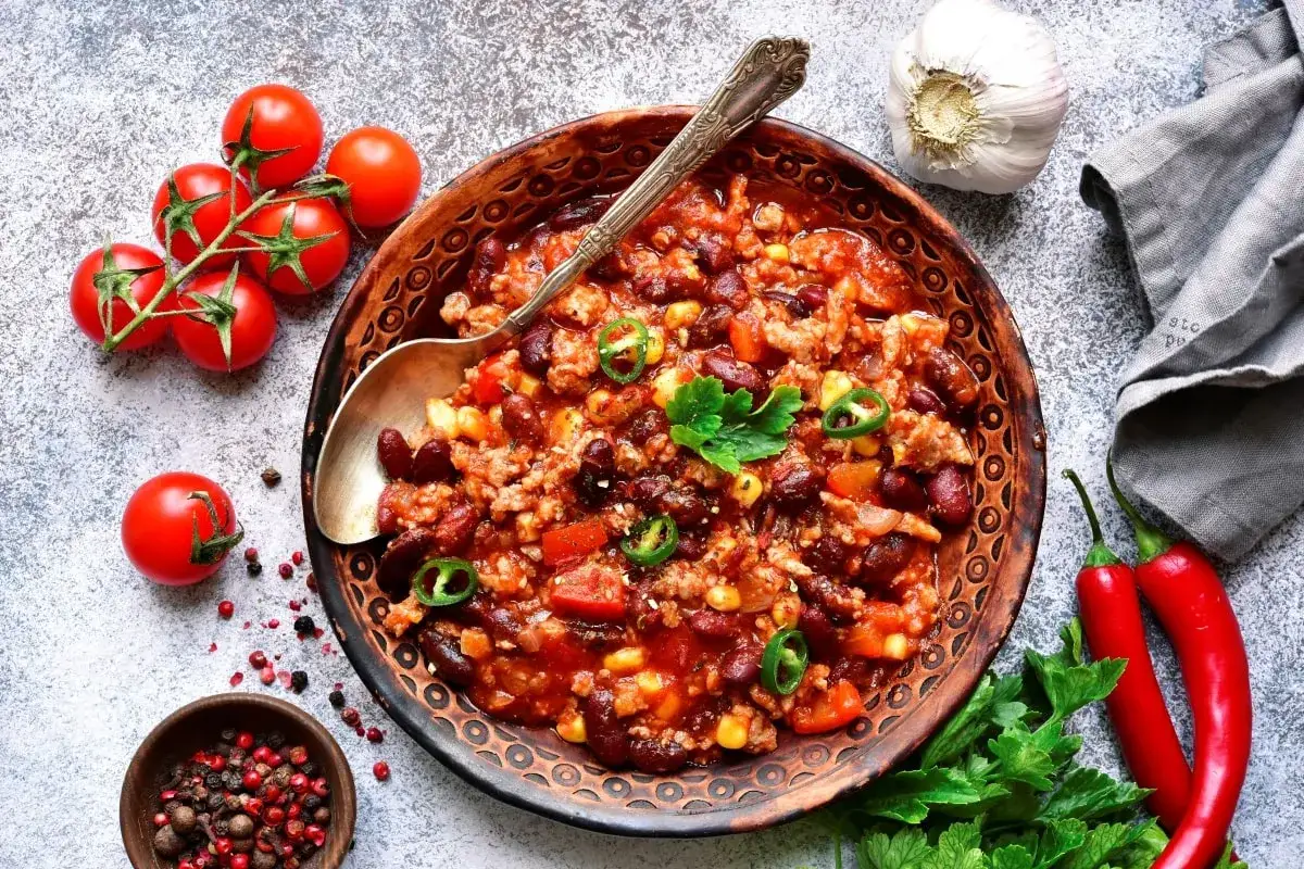 Pyszne chili con carne przepis w misce, z fasolą, mięsem mielonym, kukurydzą i pomidorami. Obok czosnek, papryczki chili i świeże pomidorki.
