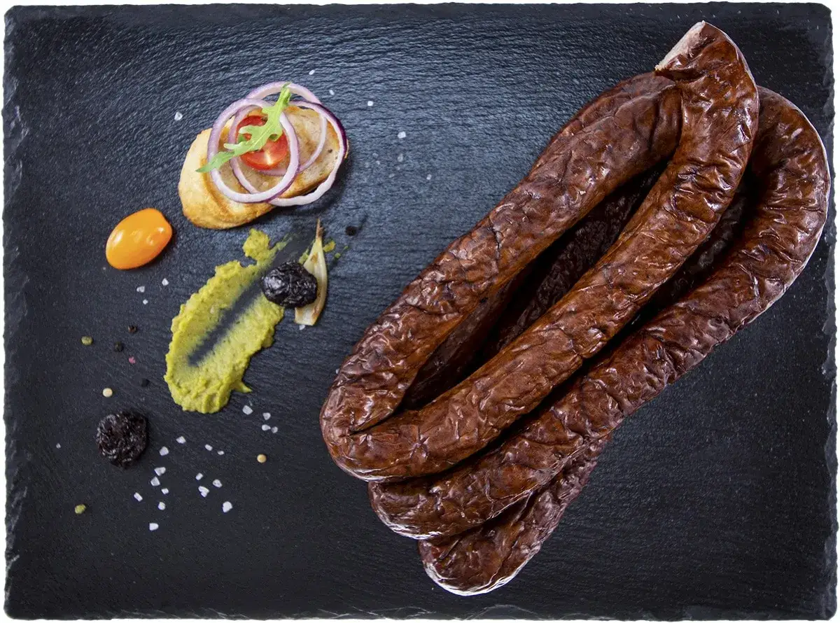 Kiełbasa z dzika przepis na 10 kg. Wędzona, ciemnobrązowa, ułożona na czarnej desce, obok dodatki: pieczywo z cebulą i rukolą, pasta, suszone śliwki, pomidorek.