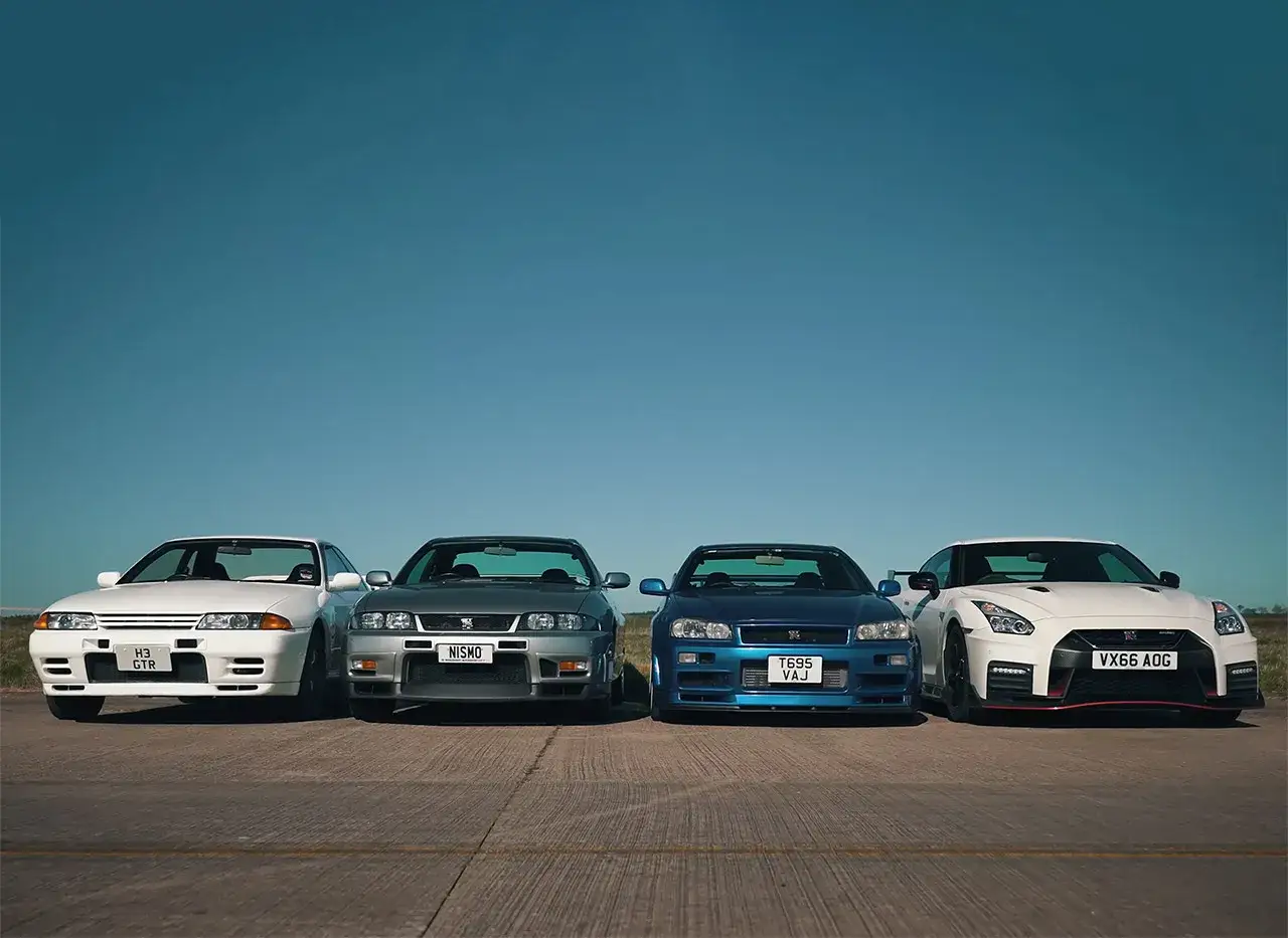Cztery ikony motoryzacji: biały, szary, niebieski i biały Nissan Skyline, stojące obok siebie na tle błękitnego nieba.