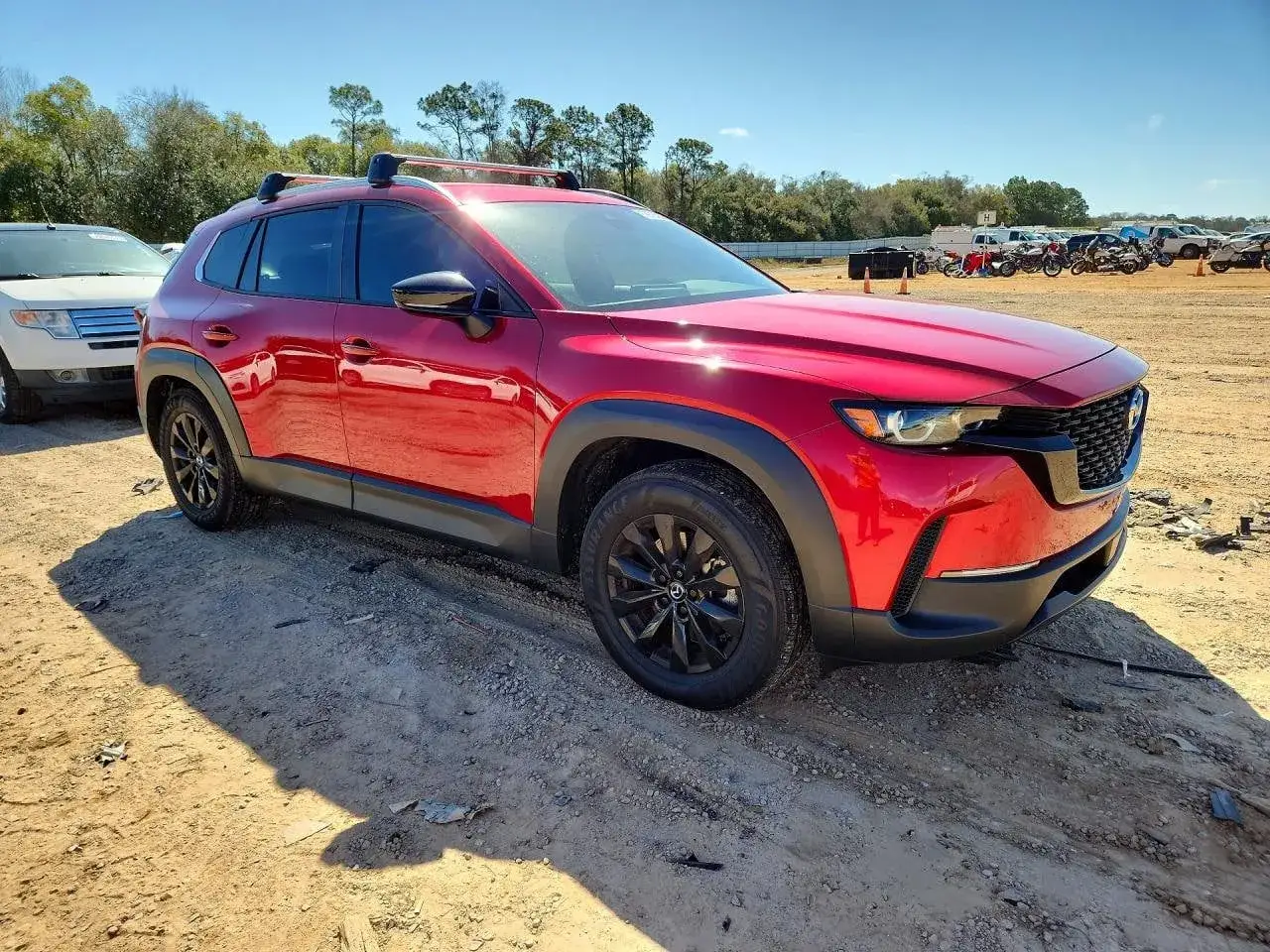 Czerwona Mazda CX-50 z czarnymi relingami dachowymi i felgami stoi na piaszczystym placu.