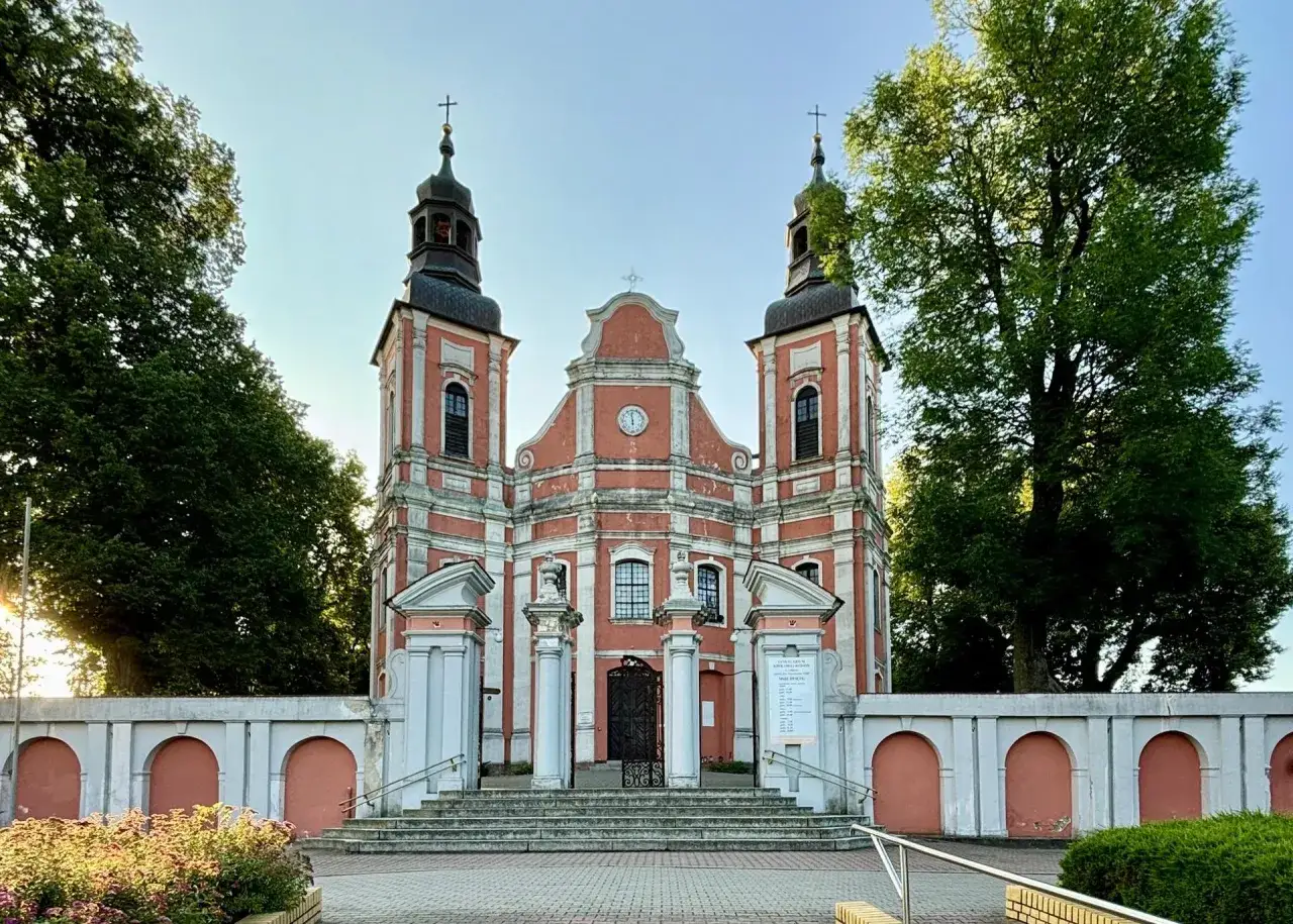 Barokowy kościół w Połajewie z dwiema wieżami i zegarem na fasadzie, otoczony murem z arkadami.