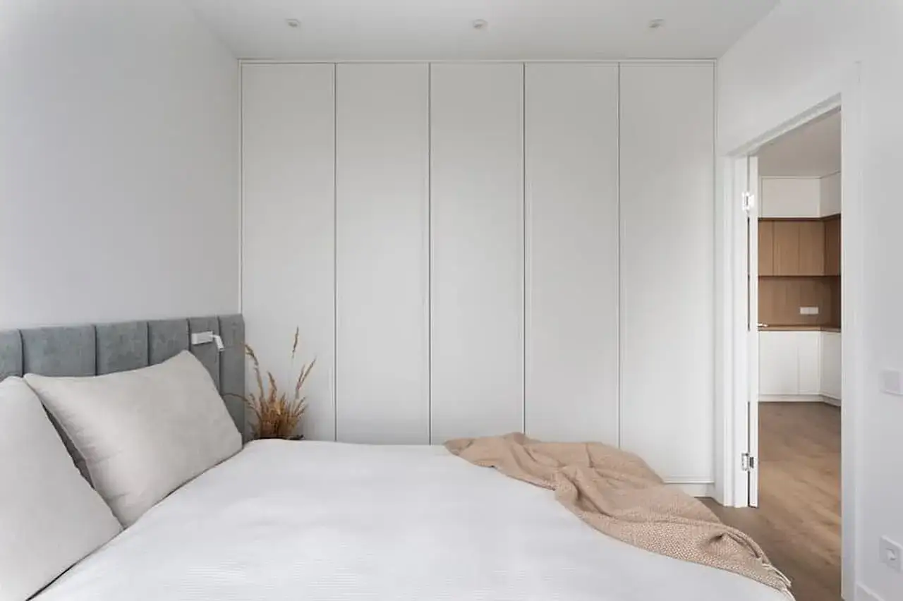 Minimalistyczna sypialnia z dużym łóżkiem i pojemną szafą. Widok na kuchnię przez otwarte drzwi. Idealny plan mieszkania 35m2.
