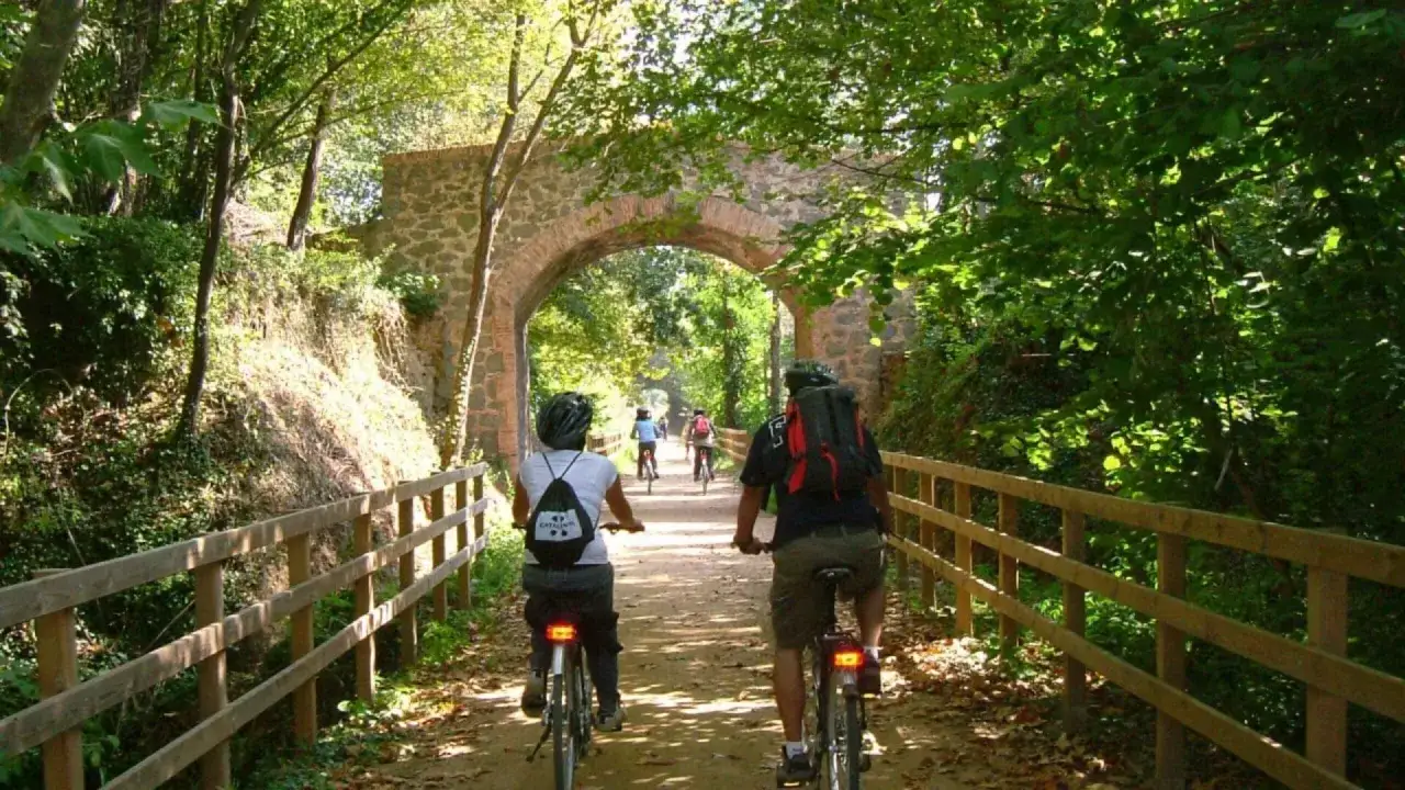 Ciclistas disfrutan de la ruta el carrilet bajo un arco de piedra, rodeados de vegetación exuberante.