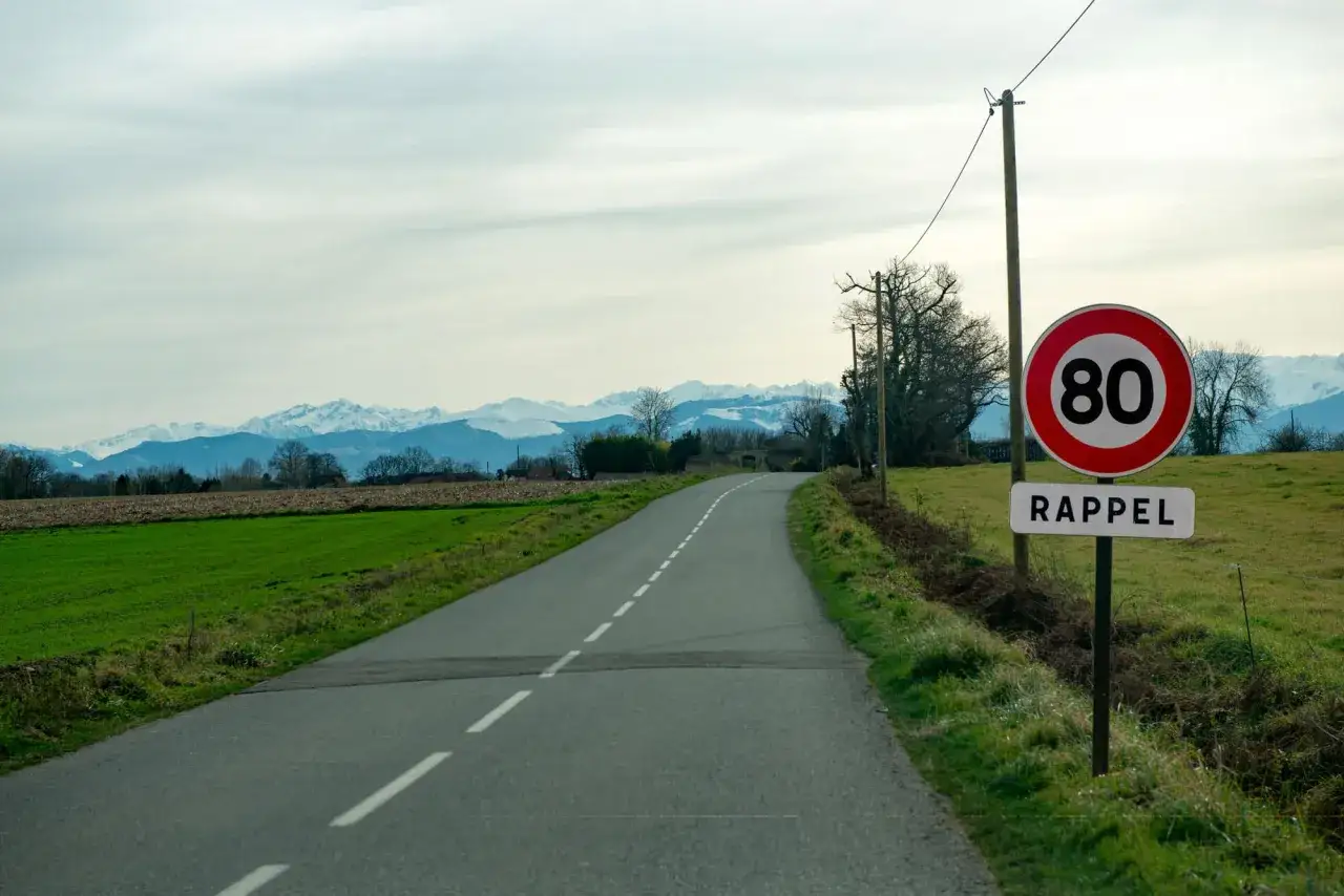 Panneau de limitation de vitesse hors agglom&eacute;ration : 80 km/h. Route de campagne avec montagnes enneig&eacute;es en arri&egrave;re-plan.